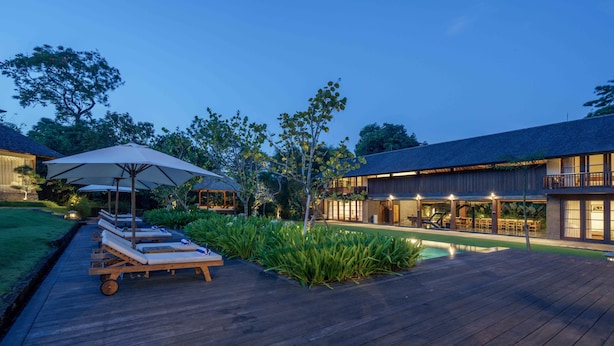 Una terraza de madera con sillones de descanso y sombrillas, una piscina y una casa moderna con amplios ventanales y mobiliario exterior.