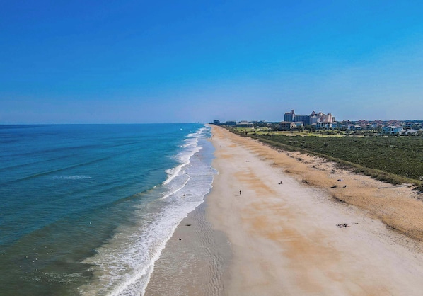 Una playa con una extensa franja de arena, un cielo azul despejado y edificios al fondo.