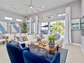 Amplia sala de estar con un ventilador de techo moderno, un conjunto de sofás, una mesa de centro con objetos decorativos y grandes ventanas que ofrecen vistas al exterior.