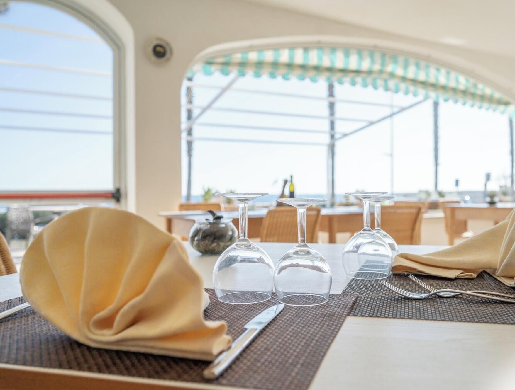 A dining table set with a folded napkin, cutlery, and wine glasses, with a view of the outdoors through large windows.
