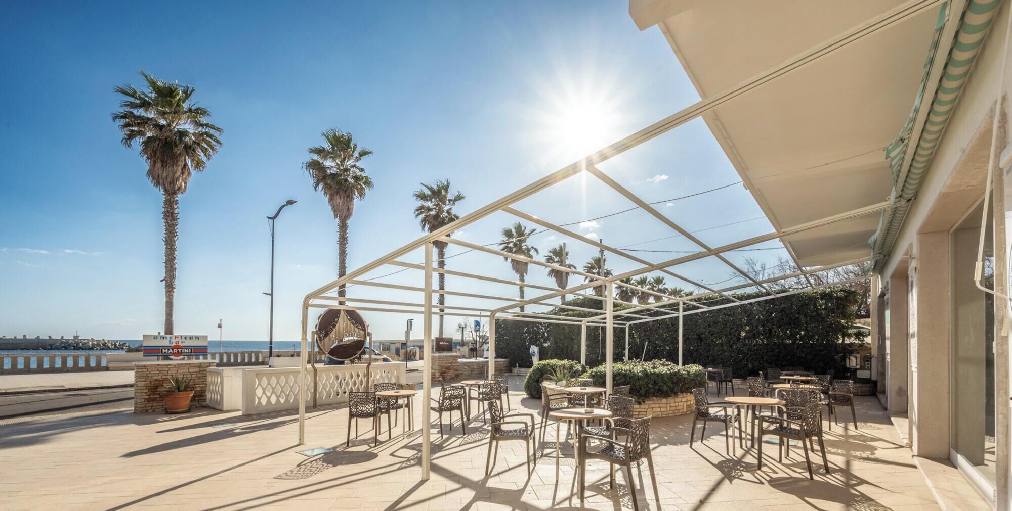 An outdoor patio with tables and chairs, palm trees, and a view of the sea.