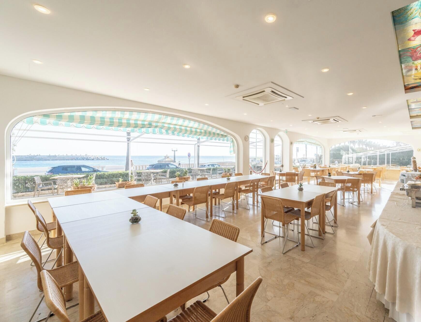 Un comedor amplio con varias mesas y sillas, ventanas grandes con vistas al mar y un techo alto con iluminación empotrada.