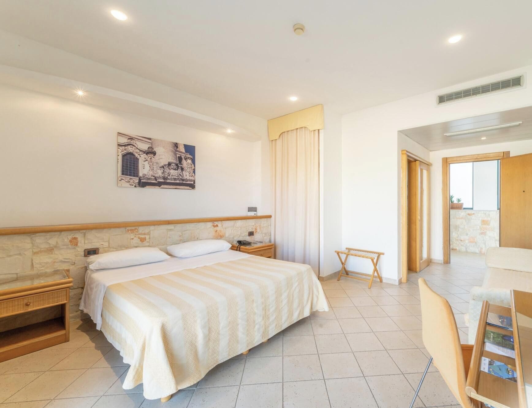 Une chambre d’hôtel avec un grand lit, une tête de lit en bois, une petite table de chevet, une commode, un tableau accroché au mur, une fenêtre avec des rideaux et une vue sur l’extérieur.