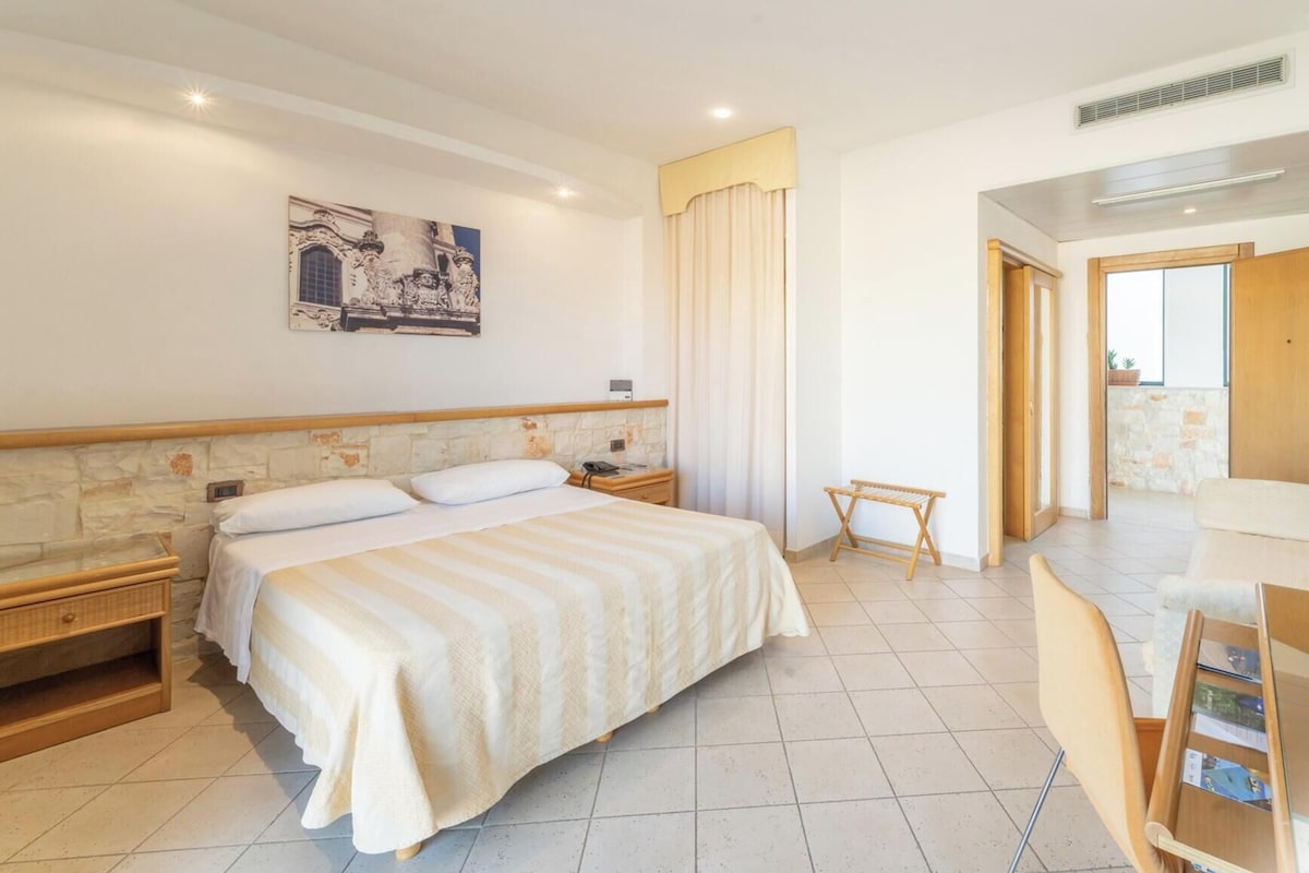 A hotel room with a large bed, a wooden headboard, a small bedside table, a nightstand, a wall-mounted painting, a window with curtains, and a view of the outside.