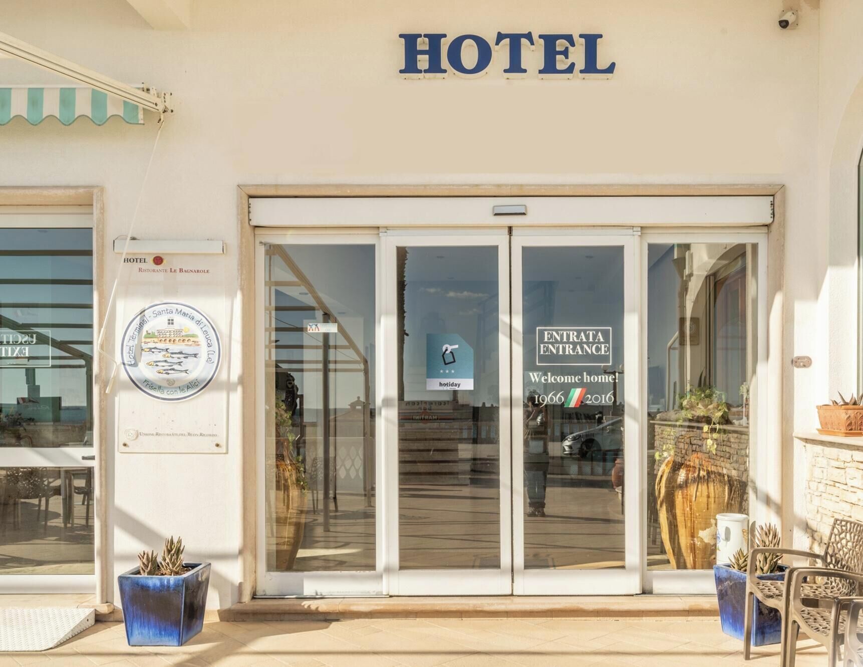 Entrada de hotel com placa indicando 'ENTRATA ENTRANCE' e mensagem de boas-vindas pelos anos de 1966 a 2016.