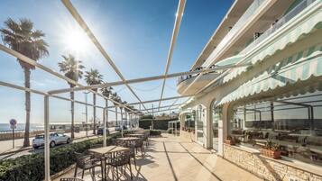 An outdoor cafe with tables and chairs, overlooking a beach and the sea.