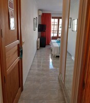 Un pasillo de hotel con una puerta de madera, un radiador, un televisor y dos camas con cortinas rojas.