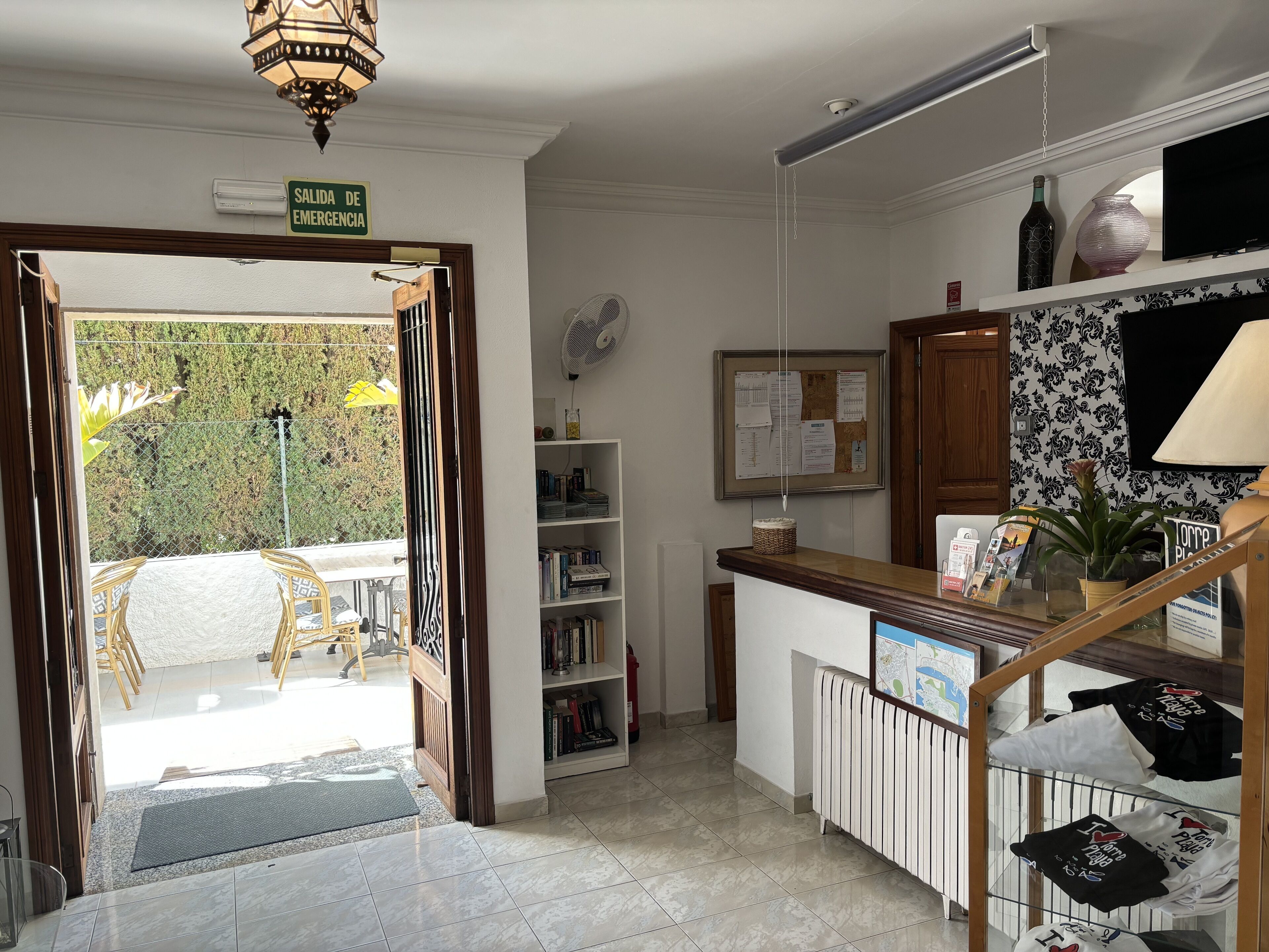 A reception area with a wooden desk, a bulletin board, a fan, and a view of the outside.
