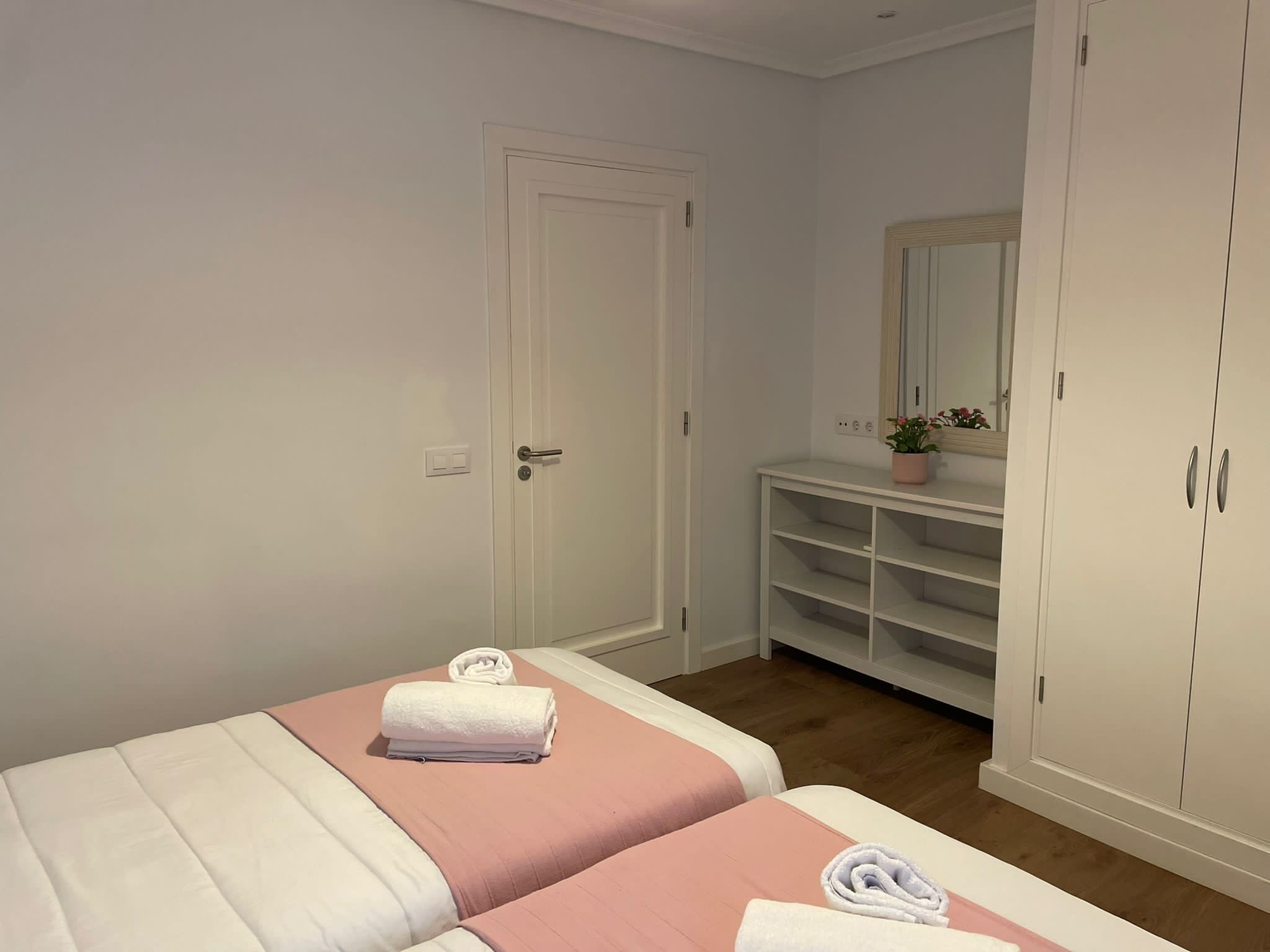 Habitación de hotel con dos camas, un armario blanco, una tocador con espejo y una pequeña planta en maceta.