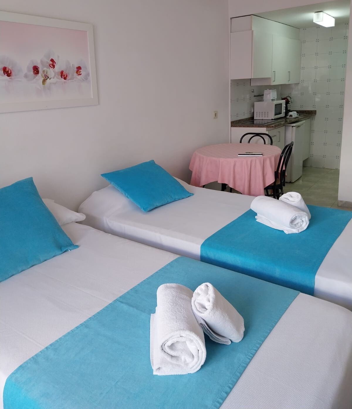A hotel room with two beds, each with blue and white bedding, a pink table, and a kitchenette in the background.