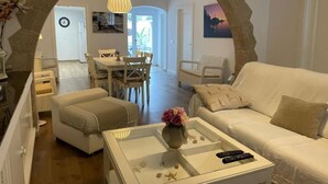 Ein Wohnzimmer mit einem weißen Sofa, einem Couchtisch mit einer Vase und Fernbedienungen sowie einem Essbereich im Hintergrund.