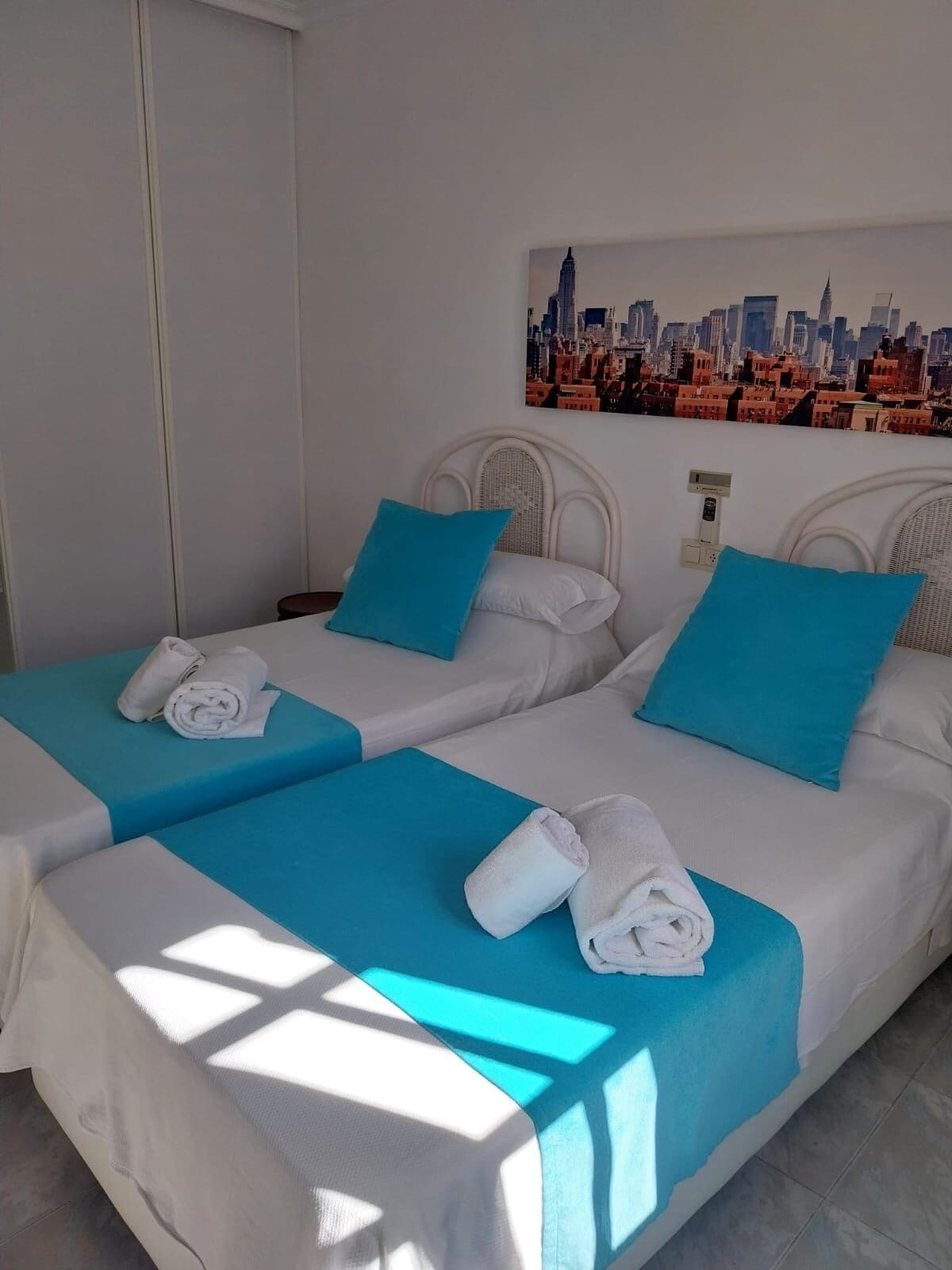 A hotel room with two beds, each with a white headboard, blue bedding, and a cityscape mural on the wall.