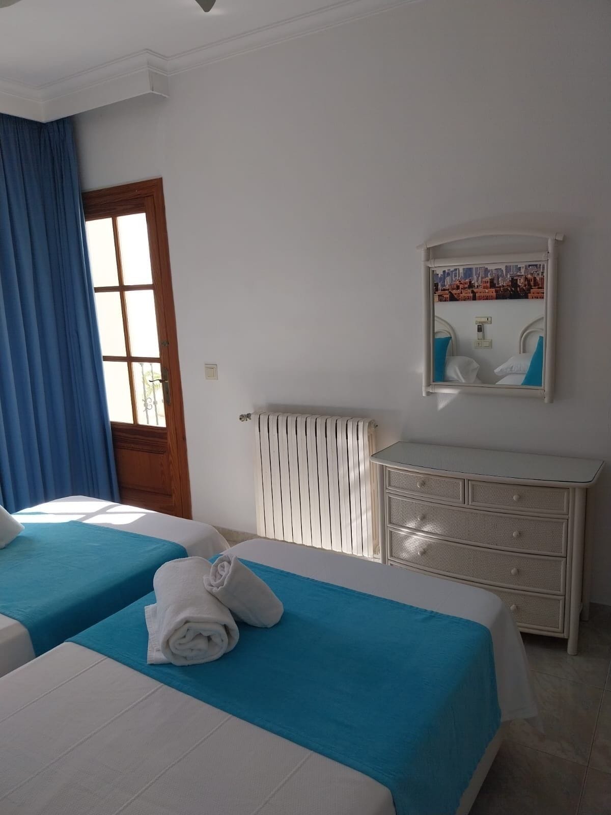 A hotel room with two beds, a dresser, a radiator, and a window with curtains.