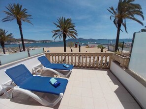 Ein Balkon mit zwei Liegestühlen, einem Fliesenboden, einem Geländer und Blick auf das Meer und die Berge.