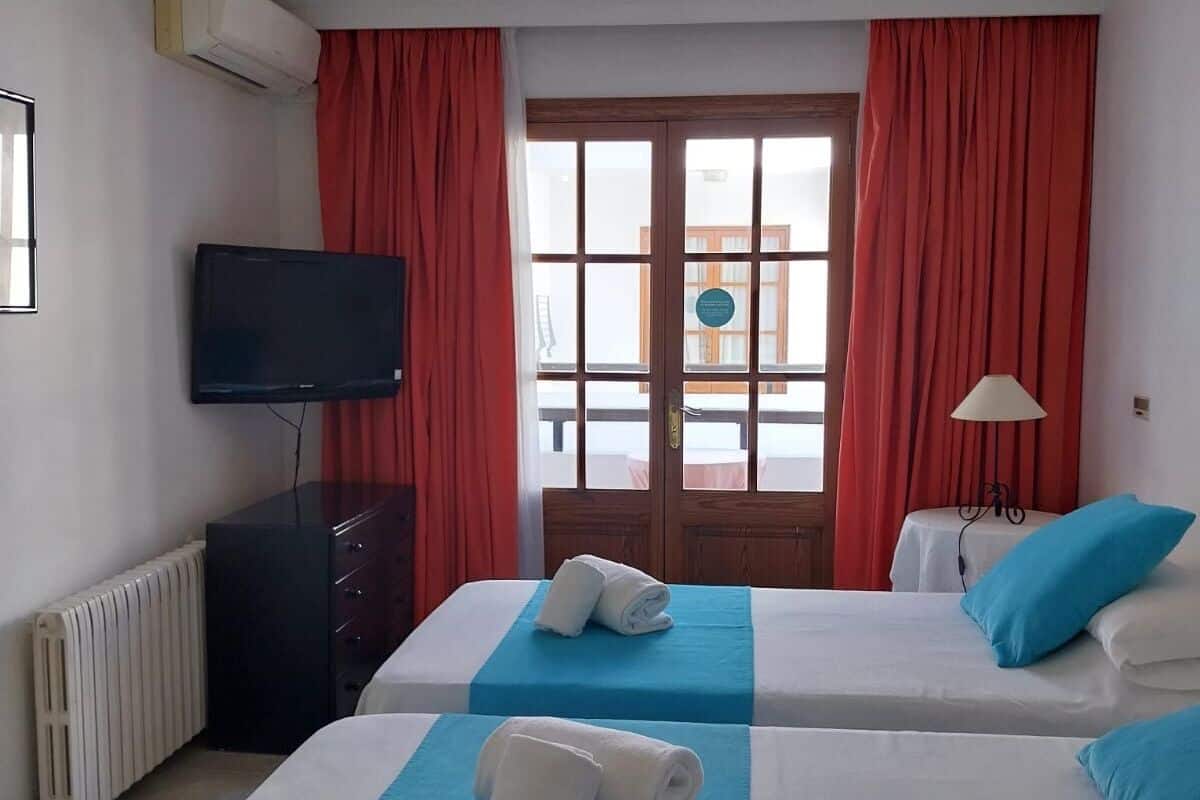 Habitación de hotel con dos camas, televisión, radiador y ventana con cortinas.