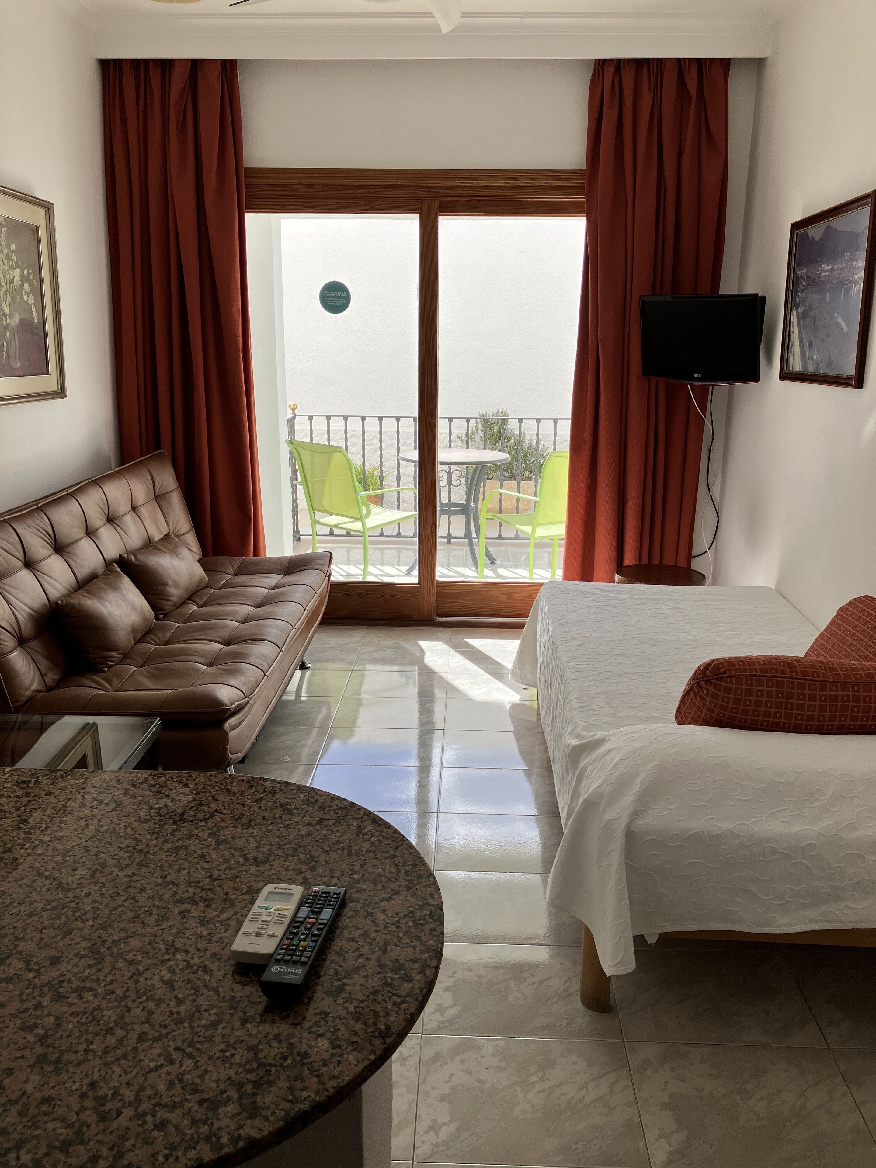 A hotel room with a brown leather sofa, a bed with white bedding, a round table with a remote control, and a balcony with green chairs and a TV.