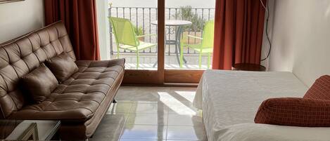 A hotel room with a brown leather sofa, a bed with white bedding, a round table with a remote control, and a balcony with green chairs and a TV.