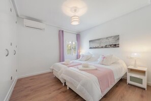Un dormitorio con dos camas, una ventana con cortinas, un aire acondicionado instalado en la pared, una mesita de noche con una lámpara y una pintura de un paisaje urbano.