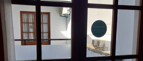 A view through a window with a balcony, featuring a round table with a pink tablecloth, a white building with a tiled roof, and a circular sign with text.