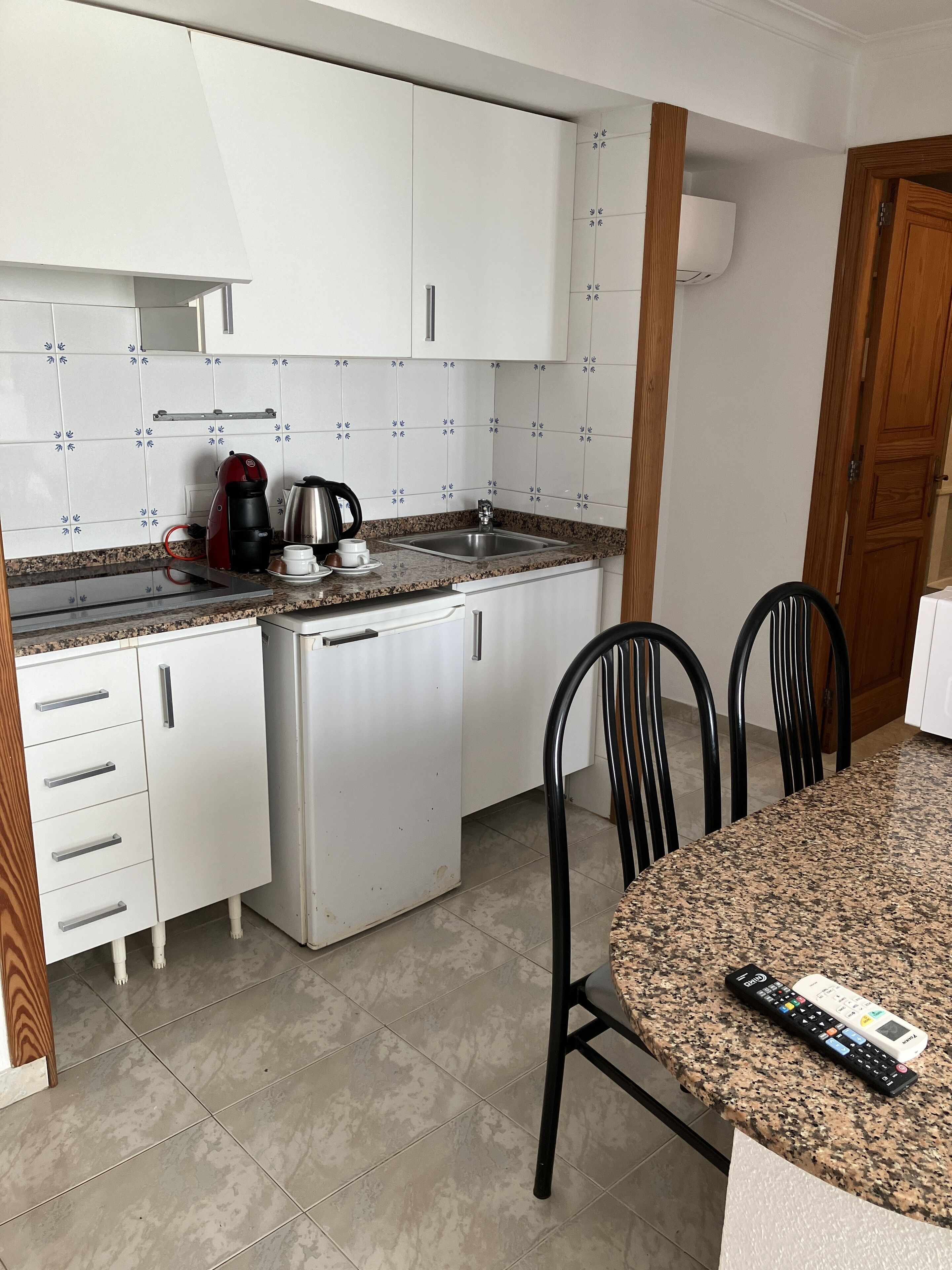 A compact kitchen with white cabinets, a granite countertop, a coffee maker, and a sink. There are two chairs at a small dining table.