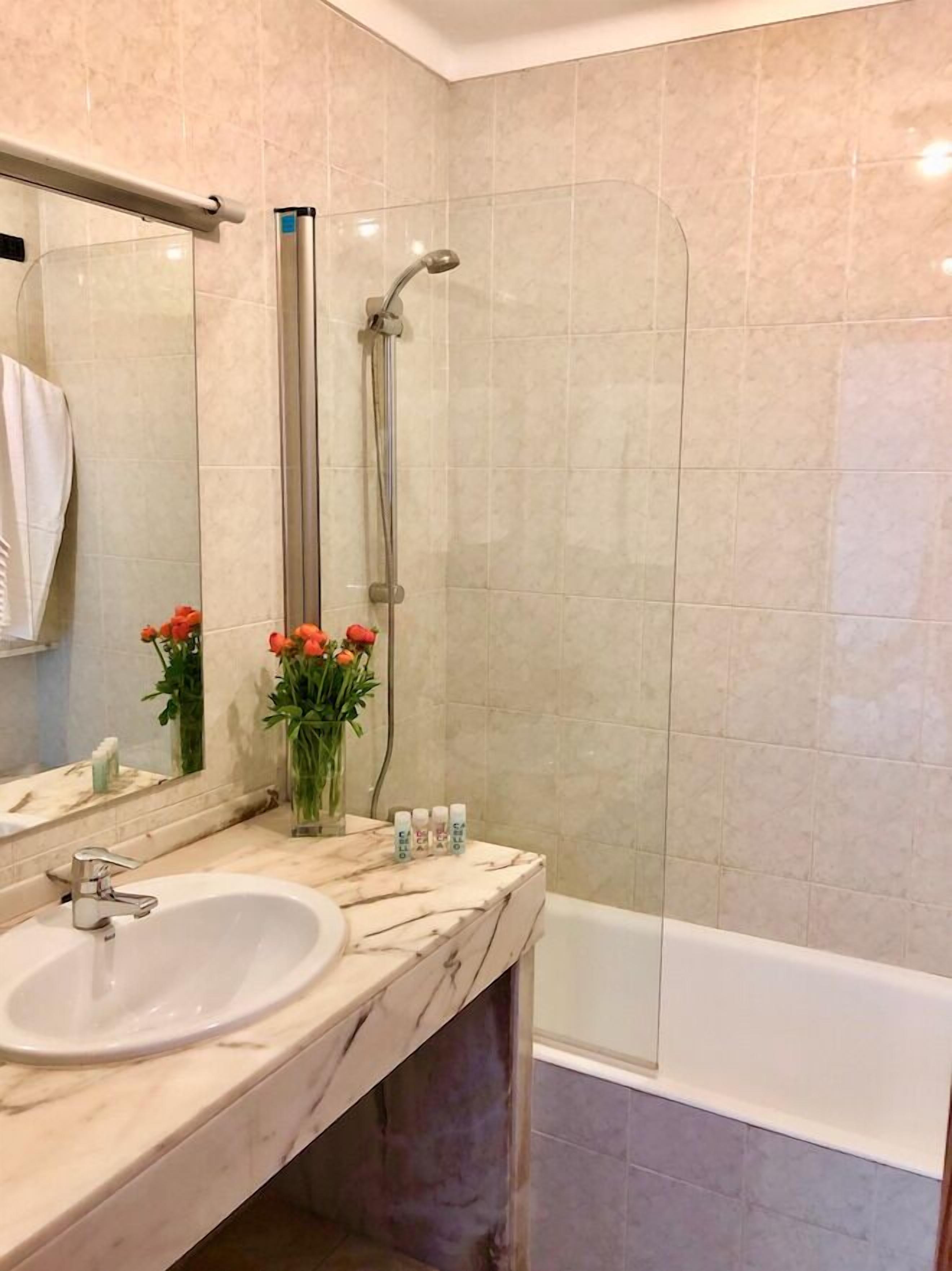 A bathroom with a marble sink, a mirror, a shower with a glass door, and a vase with flowers.