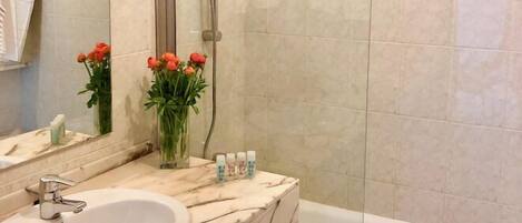 A bathroom with a marble sink, a mirror, a shower with a glass door, and a vase with flowers.