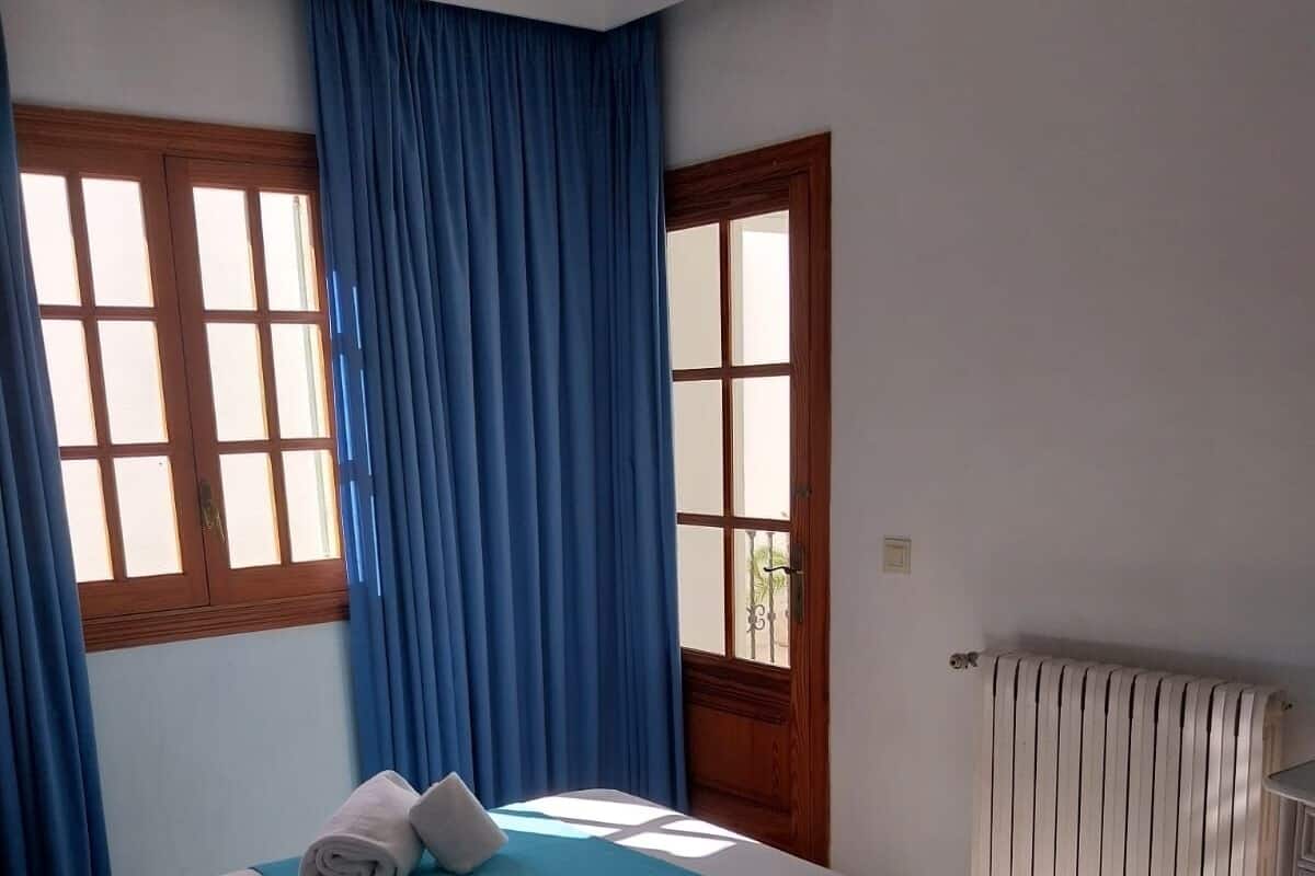 Habitación de hotel con dos camas, cortinas azules, ventanas de madera y un radiador.