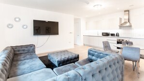 Ein modernes Wohnzimmer mit Ecksofa und Poufs, einem flachbildfernseher an der Wand und einer Küchenzeile mit weißen Schränken und Edelstahlgeräten.