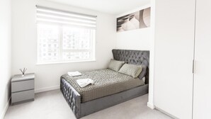 Ein Schlafzimmer mit grau gestepptem Kopfteil, einer grau-weißen Bettdecke und einem weißen Nachttisch mit Lampe.