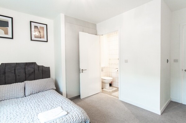 A bedroom with a bed, a wardrobe, and a bathroom visible through an open door. - Brand New Luxurious 2-bed Apartment in Barking (Barking)