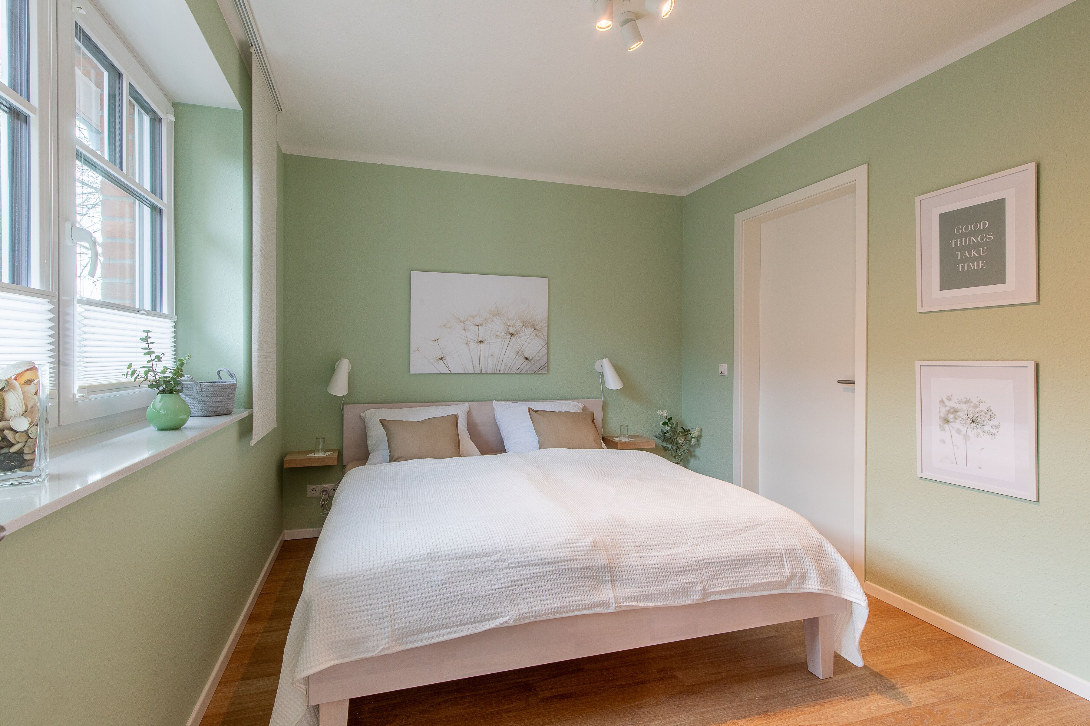 Une chambre avec un grand lit, une fenêtre avec des stores, une table de chevet avec une lampe et des tableaux encadrés au mur.