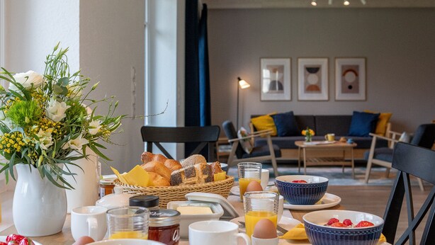 A breakfast setup with a variety of foods, including fruits, bread, and beverages, in a modern room with a sofa and artwork on the wall.