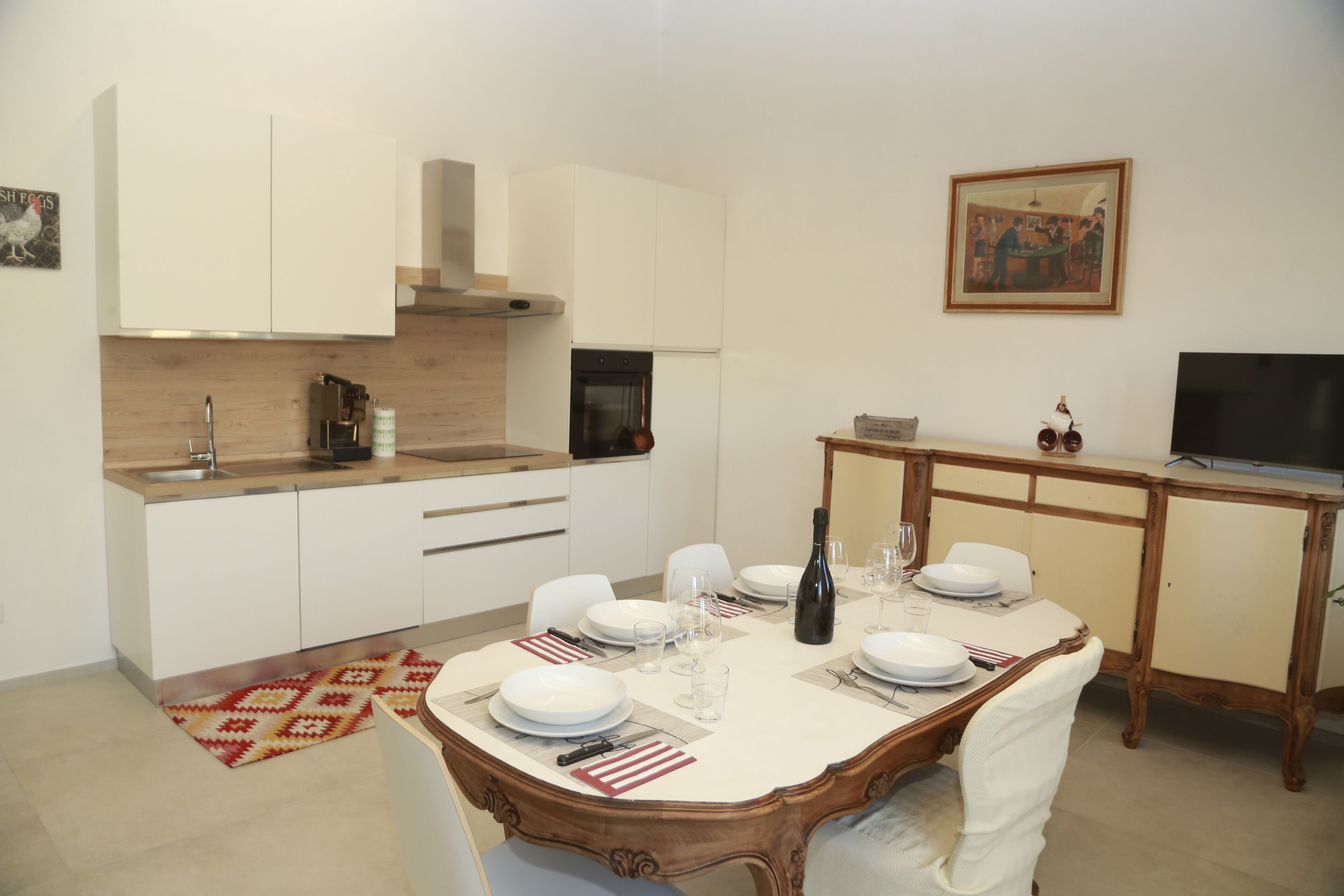 A modern kitchen with white cabinets, a built-in oven, and a dining area set for four with a table, chairs, and a TV.