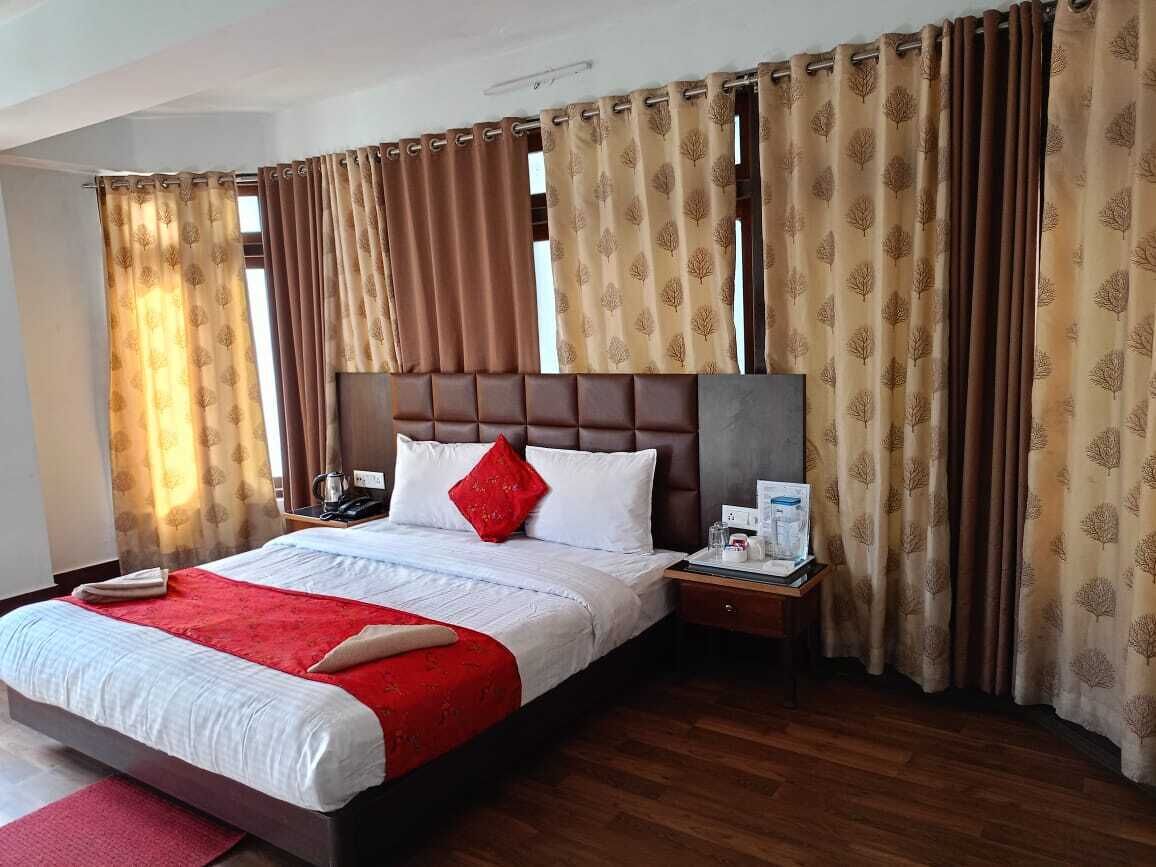 A hotel room with a large bed, a wooden headboard, a nightstand with a phone and a lamp, and curtains with a leaf pattern.