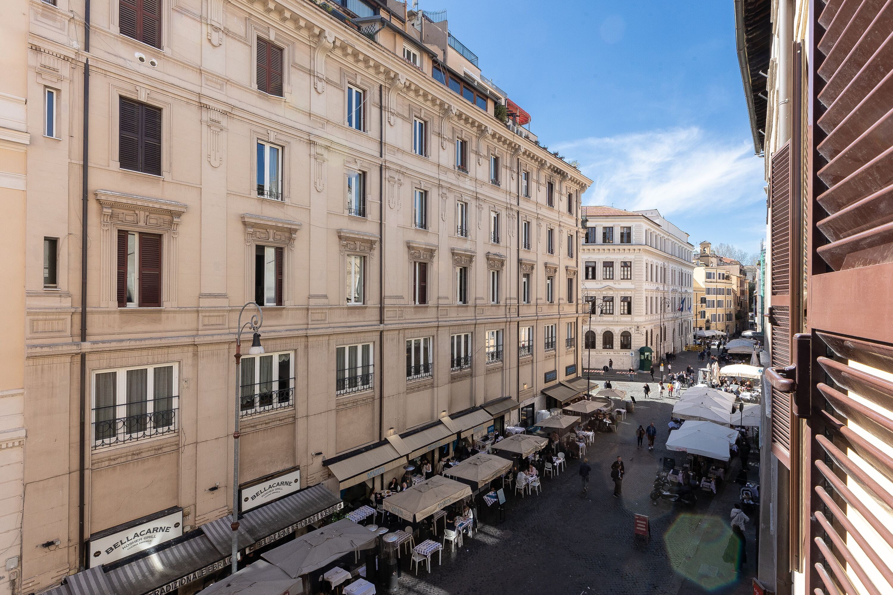 A historic building with multiple floors, large windows, and balconies. There are outdoor seating areas with tables and umbrellas, suggesting a public space or cafe.