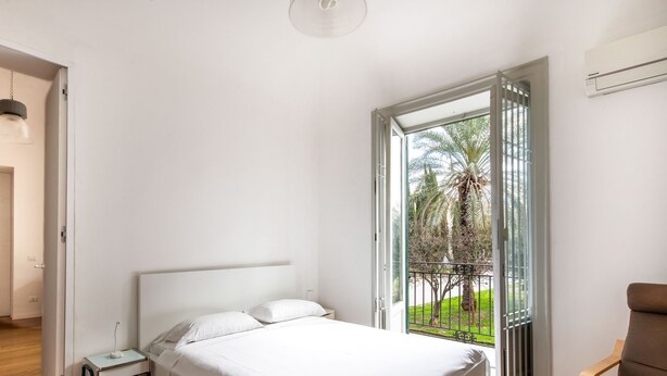 A bedroom with a bed, a bedside table, a nightstand, a window with a view of palm trees, and a door leading to another room.