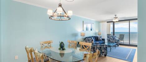 Uma sala de estar espaçosa com uma mesa redonda de vidro cercada por cadeiras de madeira, um sofá azul e poltronas estampadas. É possível ver um ventilador de teto e um lustre.