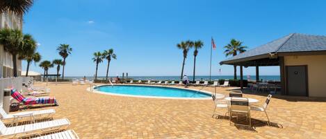 A poolside area with lounge chairs, a pool, palm trees, and a view of the ocean.