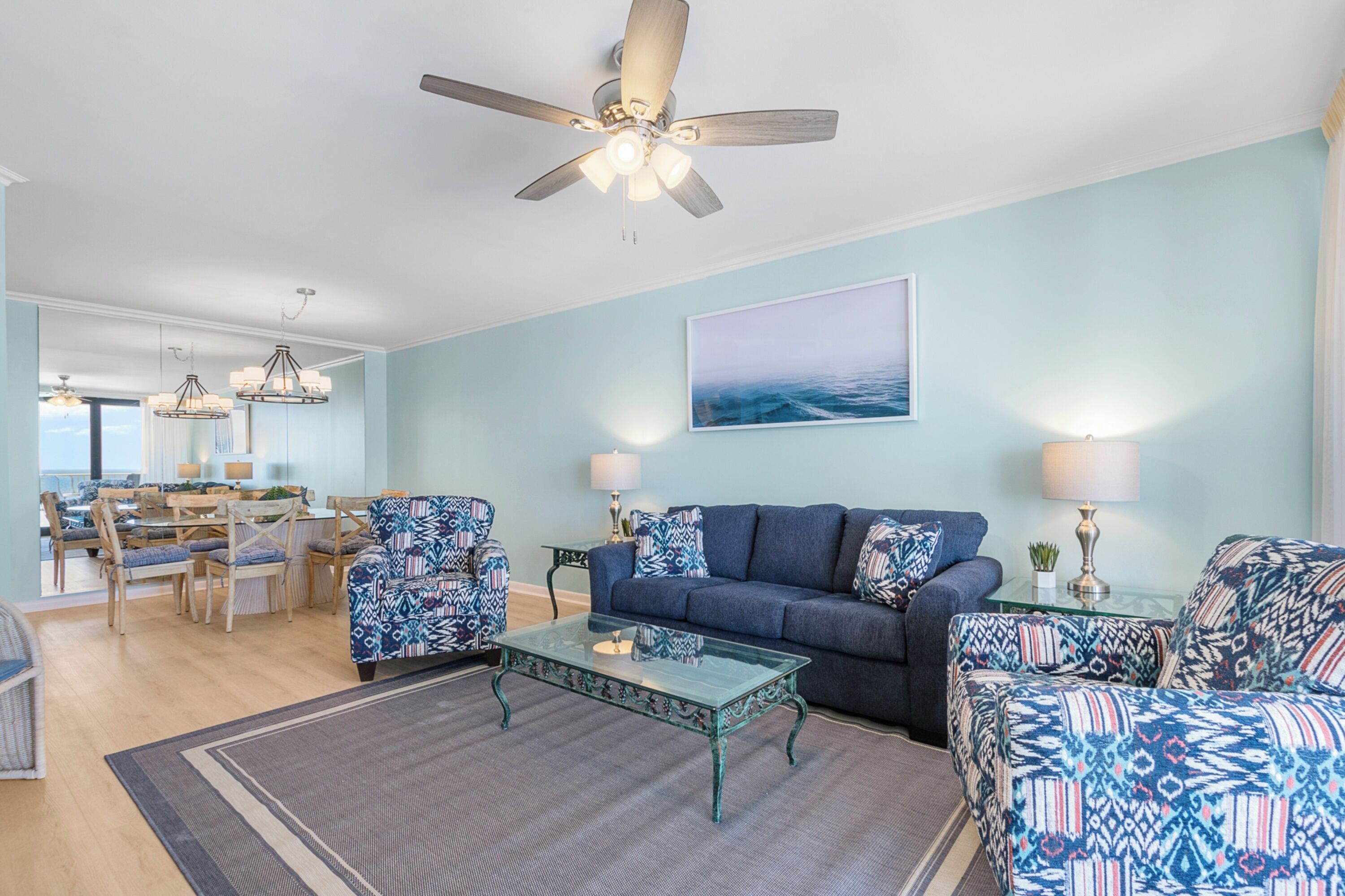 A living room with a blue sofa, patterned armchairs, a glass coffee table, and a ceiling fan.