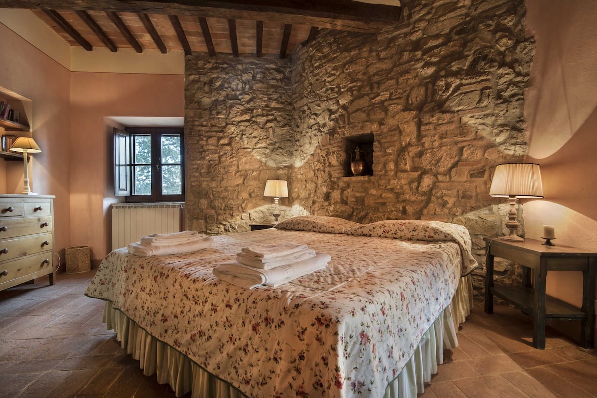 Une chambre avec un mur en pierre, un plafond en bois, un lit avec une literie à motifs floraux, une table de chevet et une fenêtre.