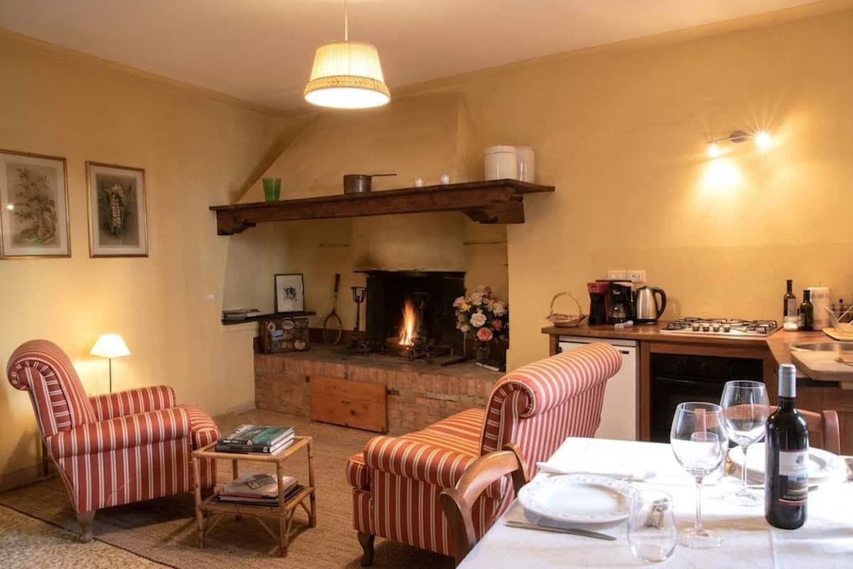 Un salon chaleureux avec des fauteuils à rayures, une cheminée, une table à manger dressée avec des verres à vin, et un coin cuisine équipé d’une cuisinière et d’une machine à café.