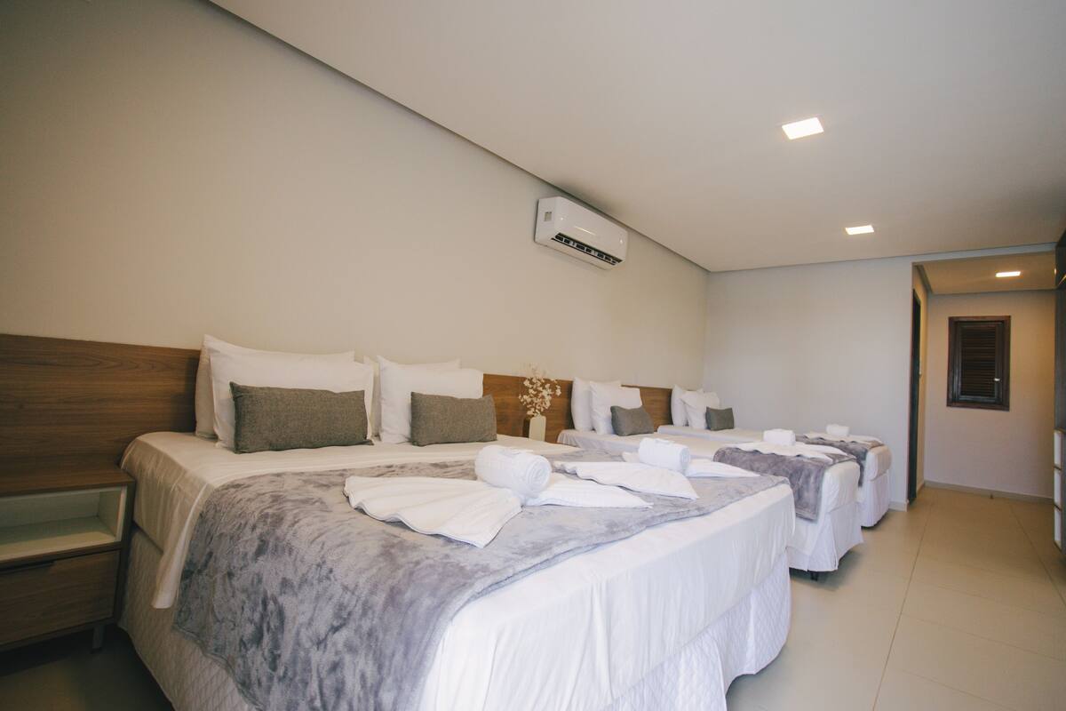 Une chambre d’hôtel avec trois lits, chacun doté de draps blancs et de couvertures grises. On y trouve une tête de lit en bois sur le côté gauche, un climatiseur fixé au mur et une petite plante accrochée au mur.