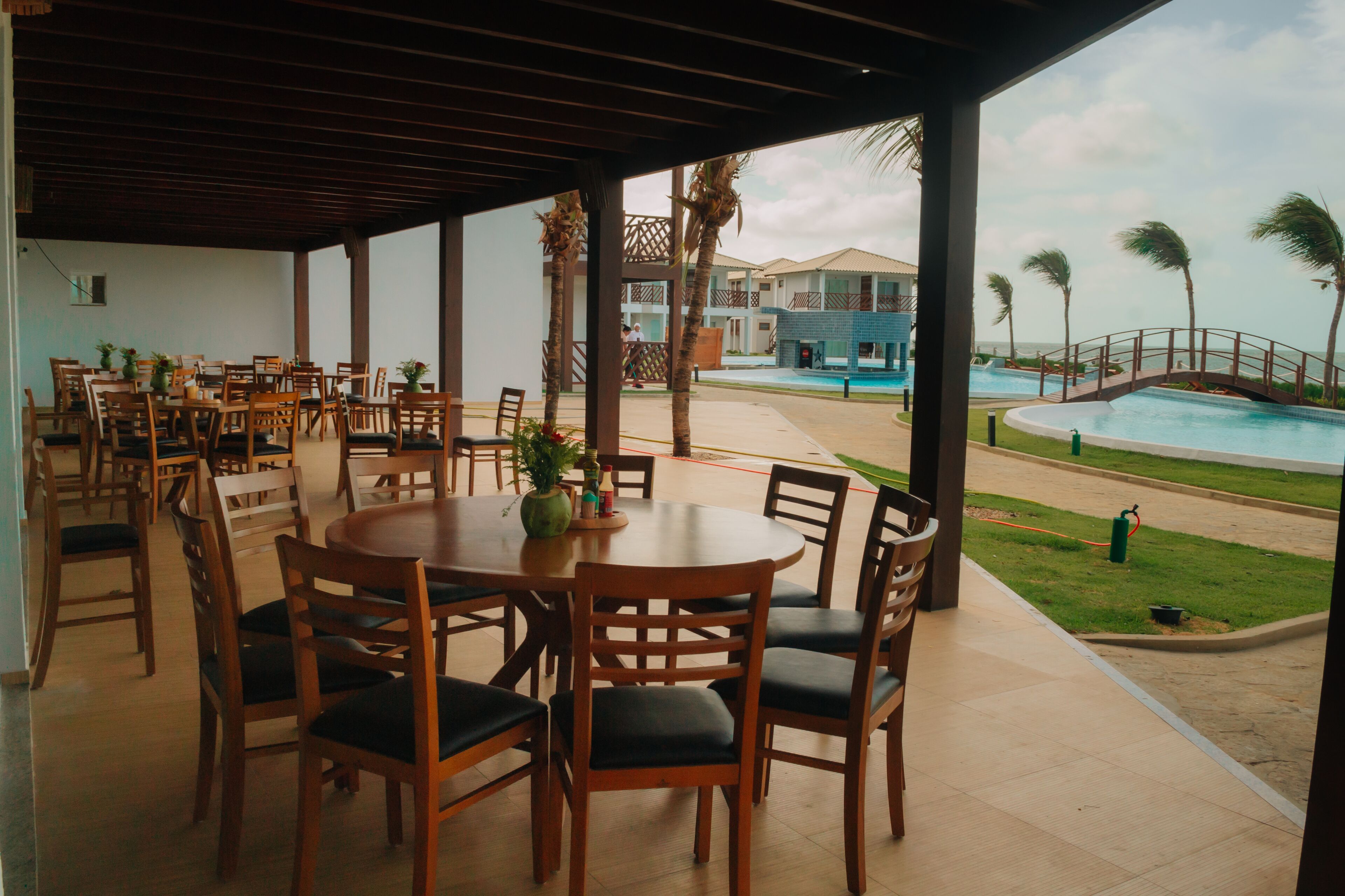 Een terras met houten tafels en stoelen, een zwembad en uitzicht op een resortgebouw.