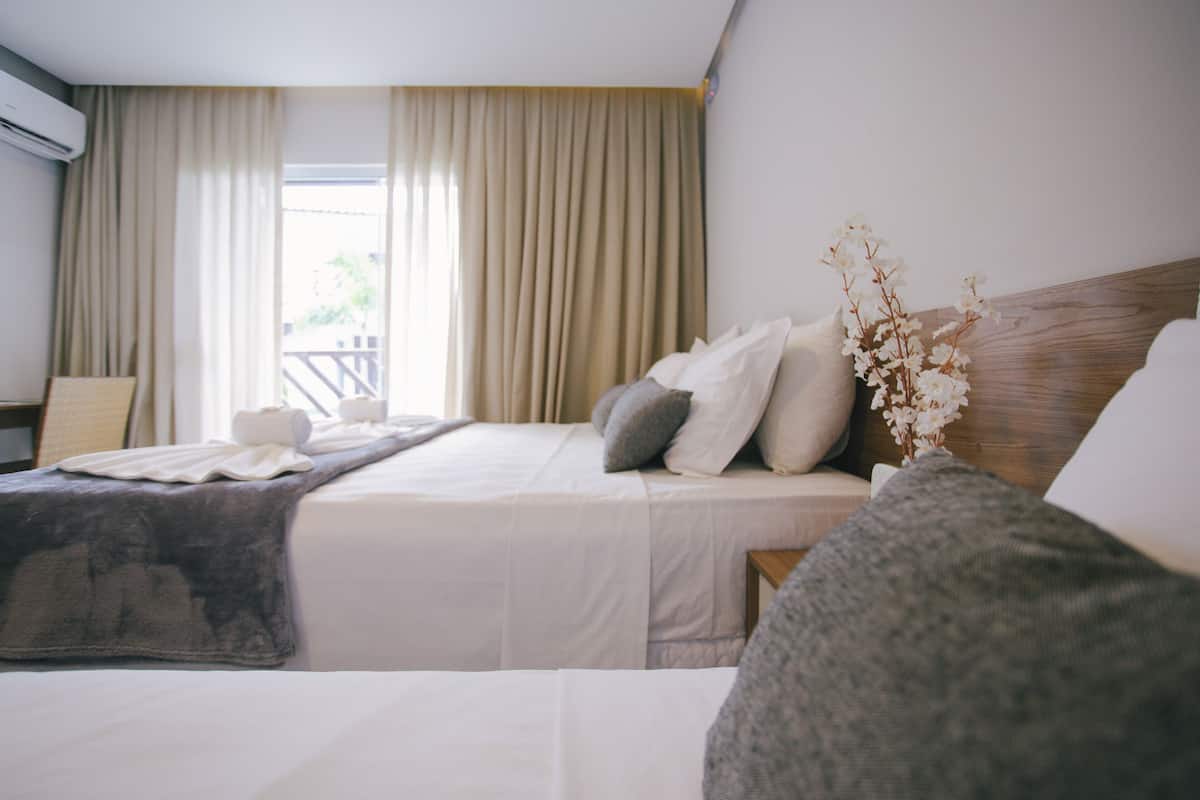 Une chambre d’hôtel avec deux lits, des rideaux beiges, une tête de lit en bois et un vase avec des fleurs blanches.
