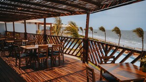 Een houten terras met tafels en stoelen, uitkijkend op een strand.