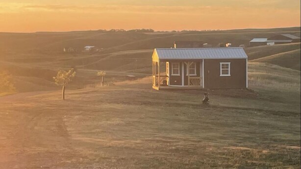 Een klein, laag huis met een veranda, omringd door open velden en heuvels.