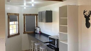 Een compacte keuken met een houten plafond, witte wanden, een ingebouwde magnetron en een koelkast. Drie krukken en een raam met zonwering.