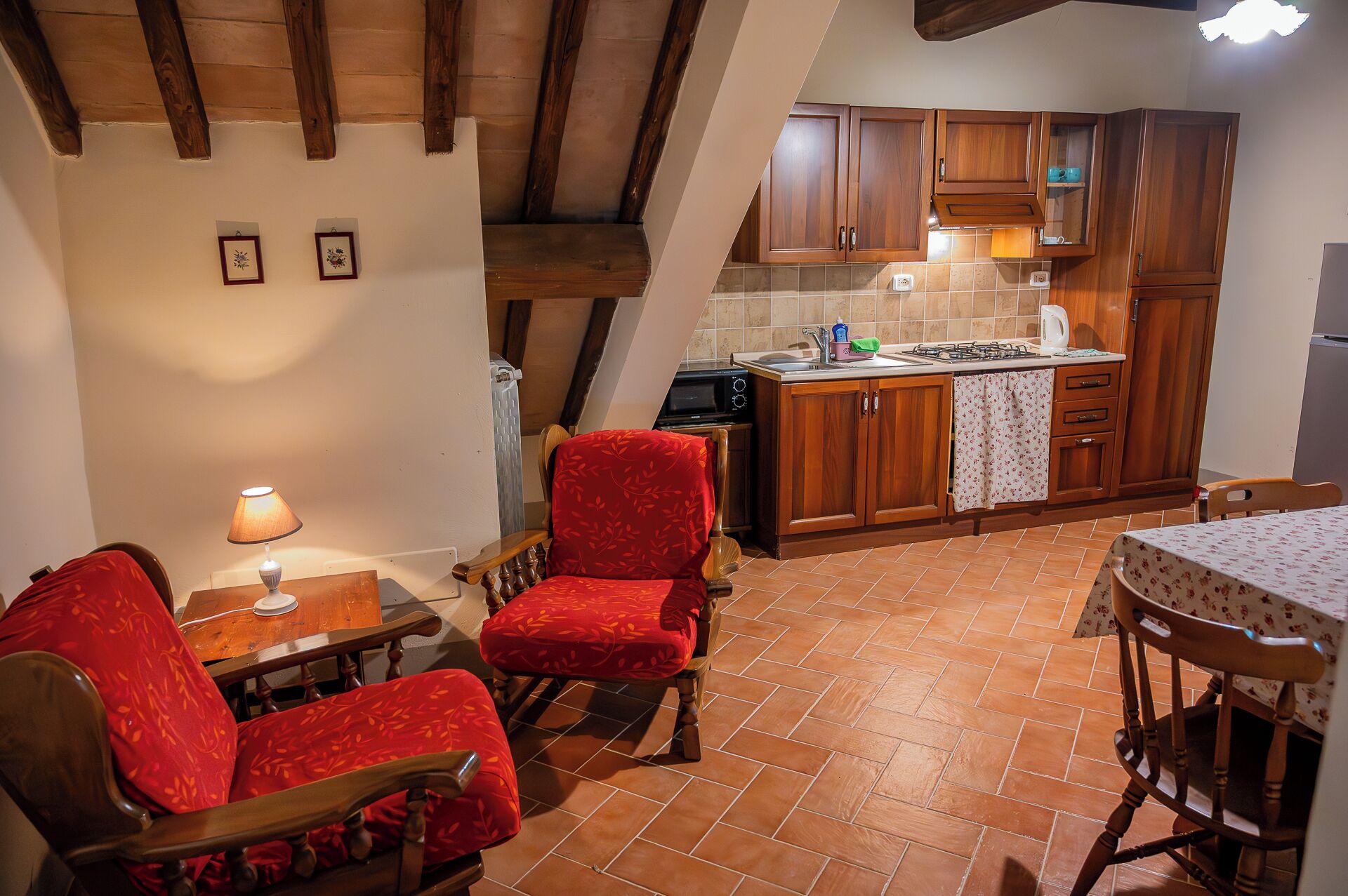 A cozy kitchen with wooden cabinets and a tiled floor, a dining area with red upholstered chairs, and a table with a lamp.