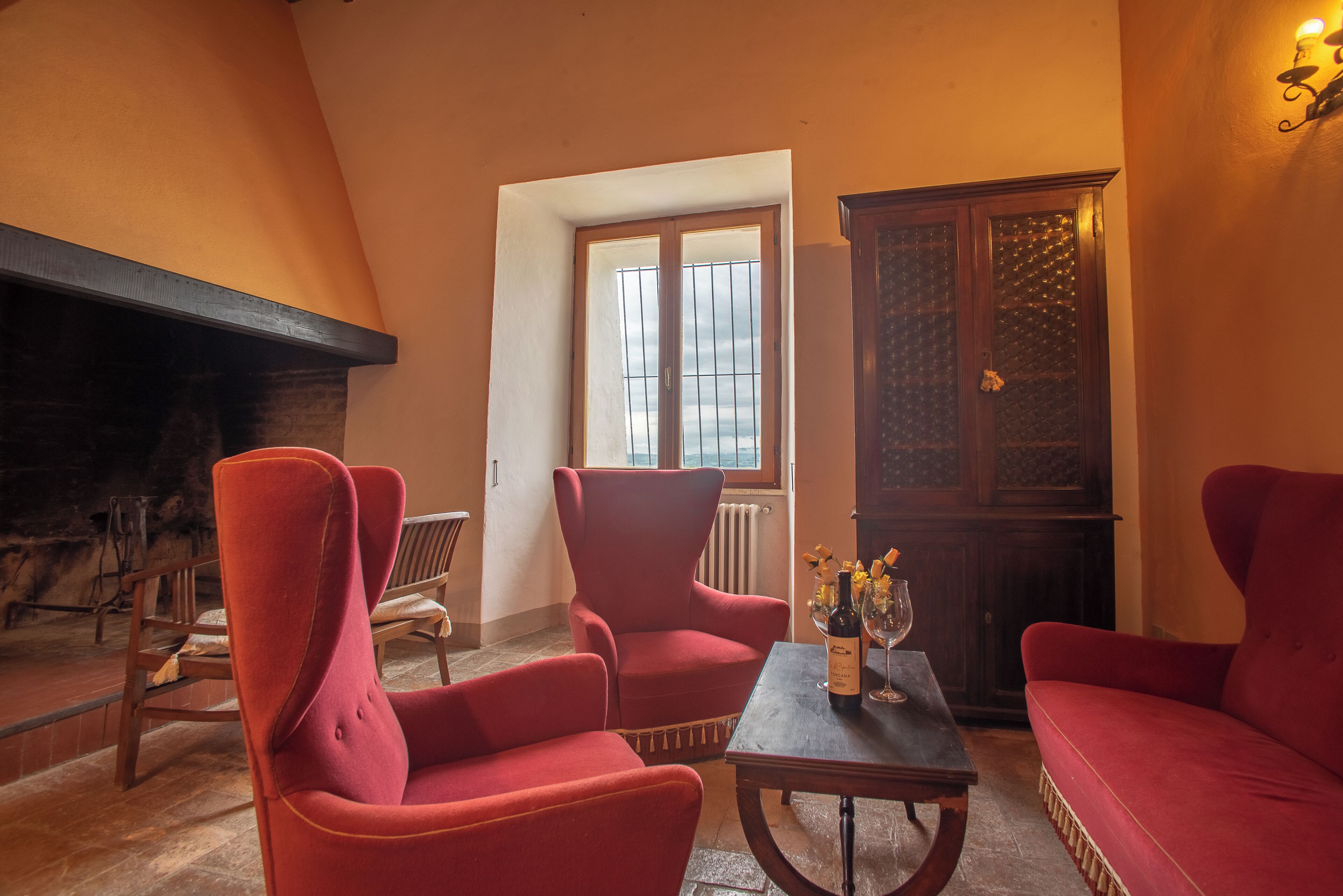 Un salon chaleureux avec une cheminée, des fauteuils rouges, une table en bois avec une bouteille et un verre, et une vue par la fenêtre.