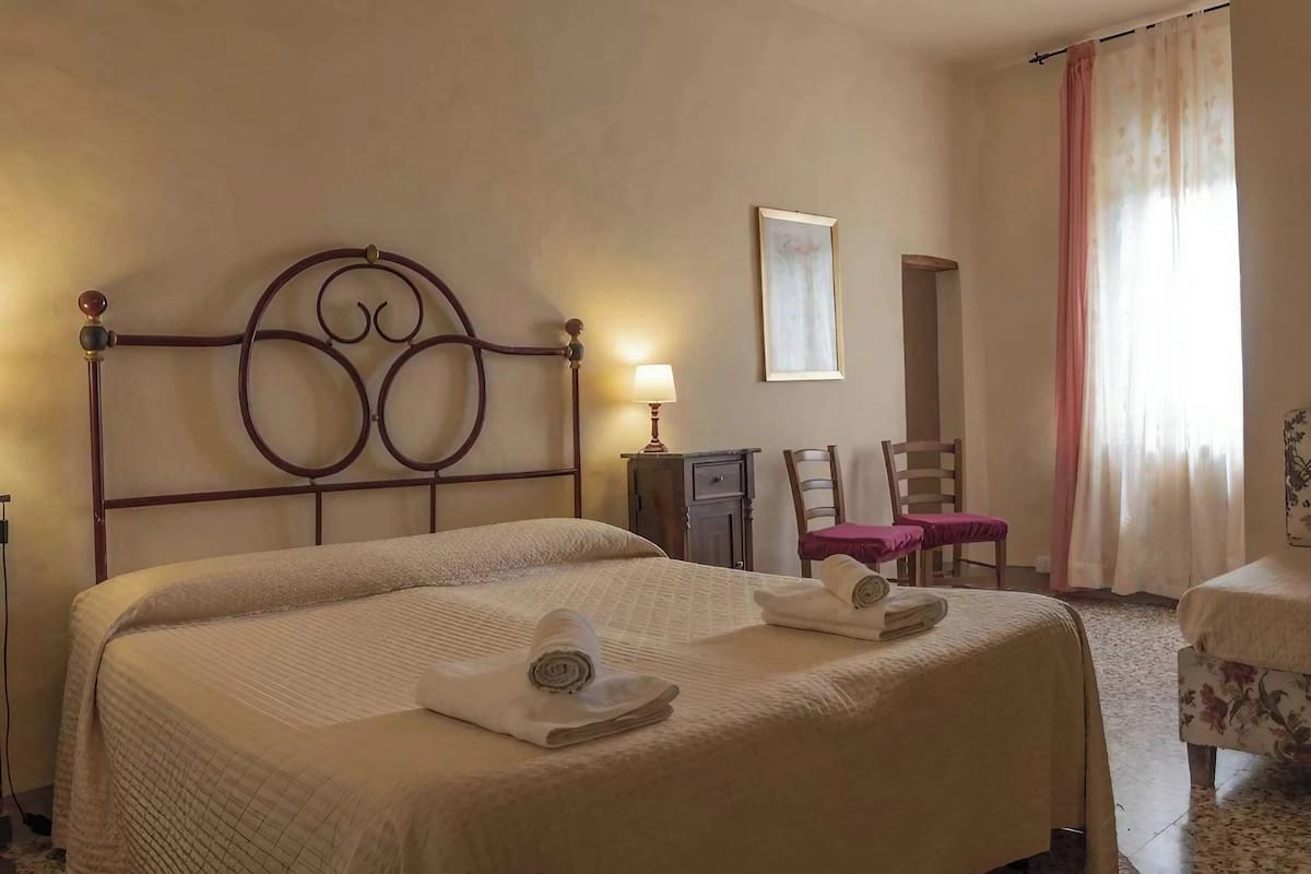 A hotel room with a double bed, a bedside table with a lamp, a chair, a small table with a pink tablecloth, and a window with curtains.